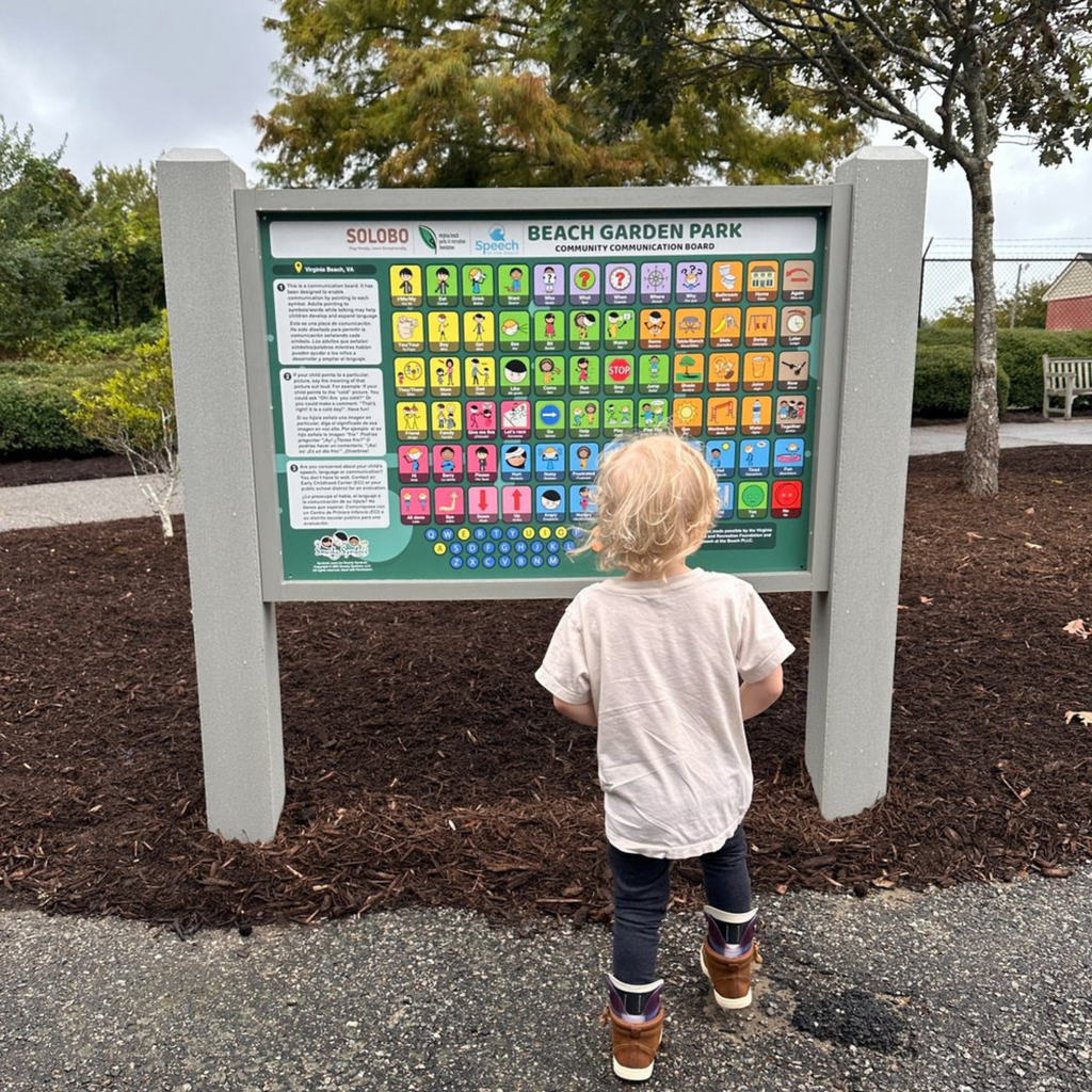 an aac board installed at a playground with a child standing in front of it