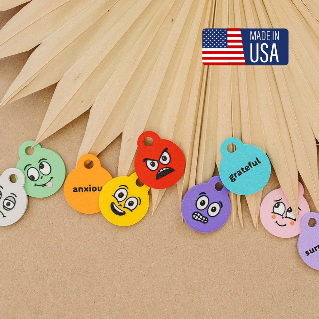A collection of colorful 3D-printed emotion keychains laid out on a wooden surface, with names like anxious, grateful, and sad printed on the back.